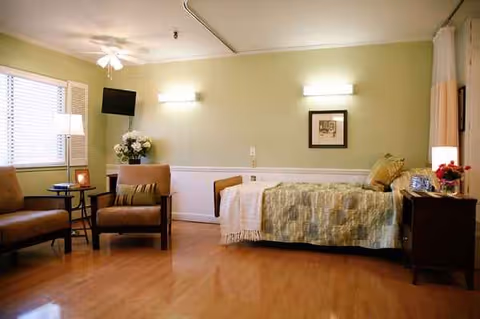 A patient bedroom with a bed, seating area, and soft lighting in a care facility.