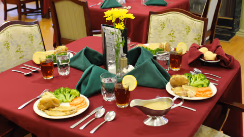 A dining table set with plates of food including broccoli, carrots, mashed potatoes, and meat. The table is covered with a red tablecloth and has green folded napkins, glasses of iced tea with lemon slices, water glasses, silverware, a small vase with yellow flowers, and a gravy boat.