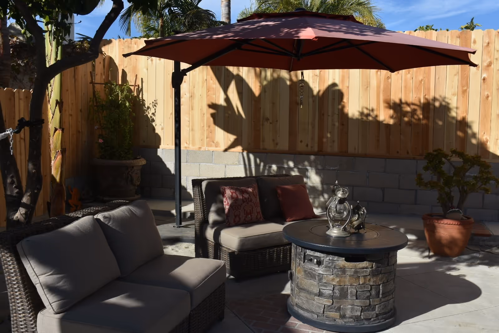 Outdoor patio area with cushioned wicker seating, a round stone fire pit table, a large red umbrella providing shade, potted plants, and a wooden fence in the background under a clear blue sky.