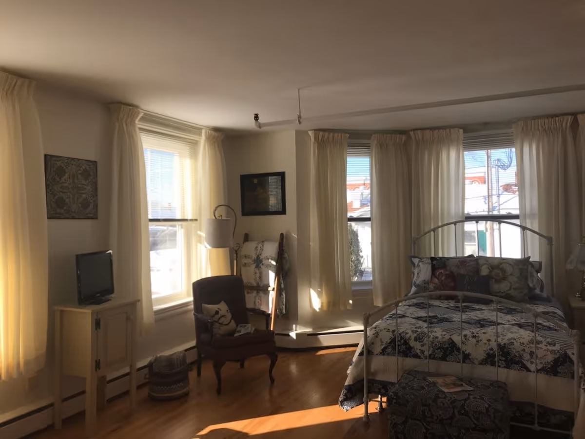 Bright bedroom with a metal-framed bed, patterned bedding, an armchair, small TV on a cabinet, and several curtained windows.