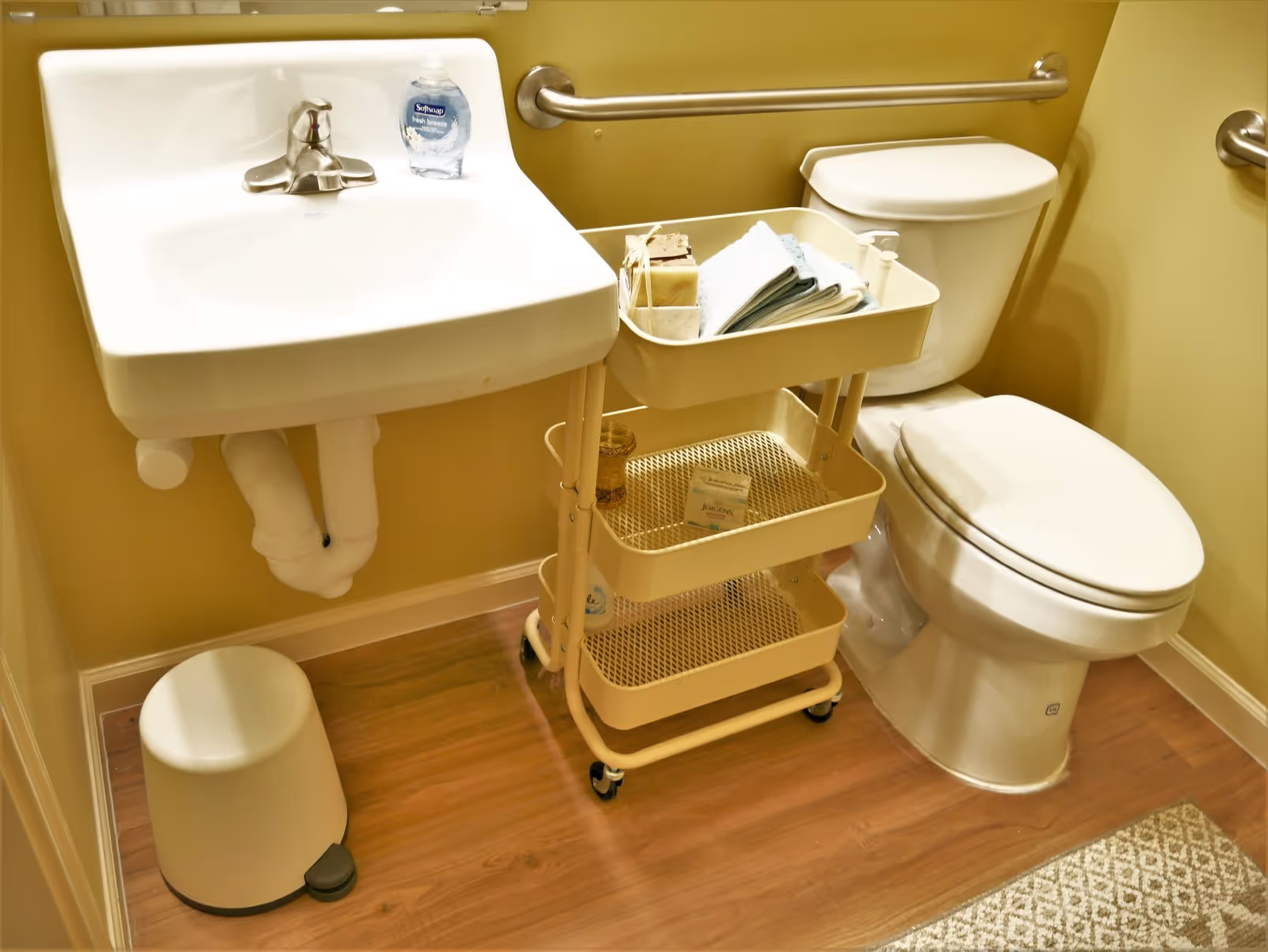 A small bathroom with a white wall-mounted sink, a bottle of Softsoap hand soap on the sink, a white toilet with a closed lid, a beige rolling cart with towels and soap, a small white trash can, and a wooden floor with a patterned rug.