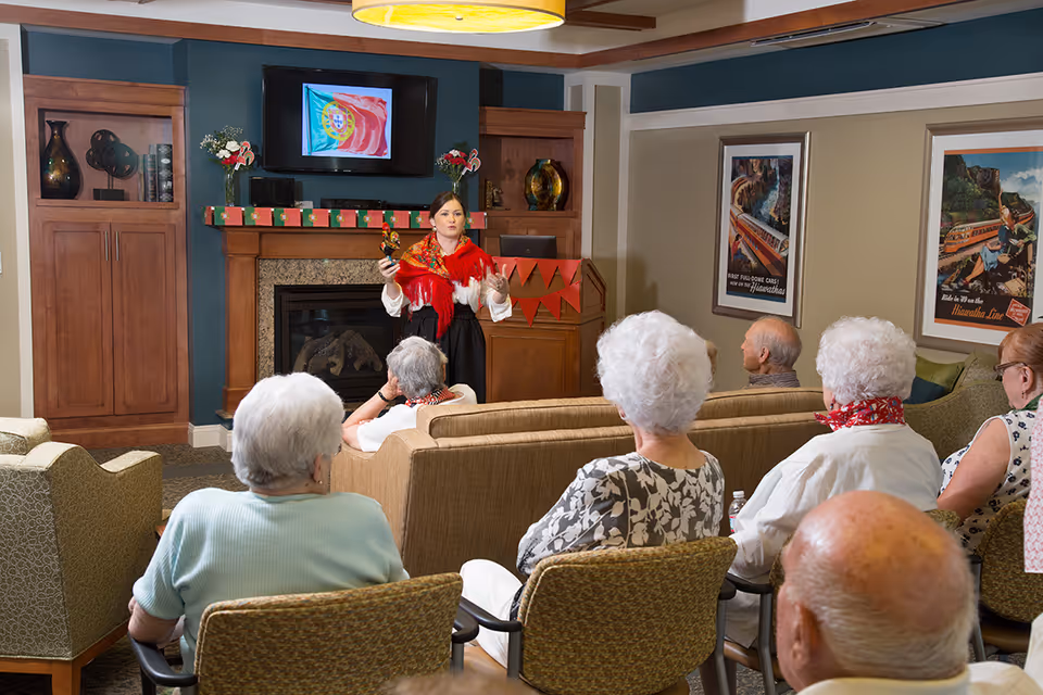 A group of elderly people seated in a living room area attentively watching a woman standing in front of a fireplace giving a presentation. The woman is dressed in traditional clothing and holding a small object. The room has wooden cabinets, framed pictures on the walls, and a TV above the fireplace displaying a flag.