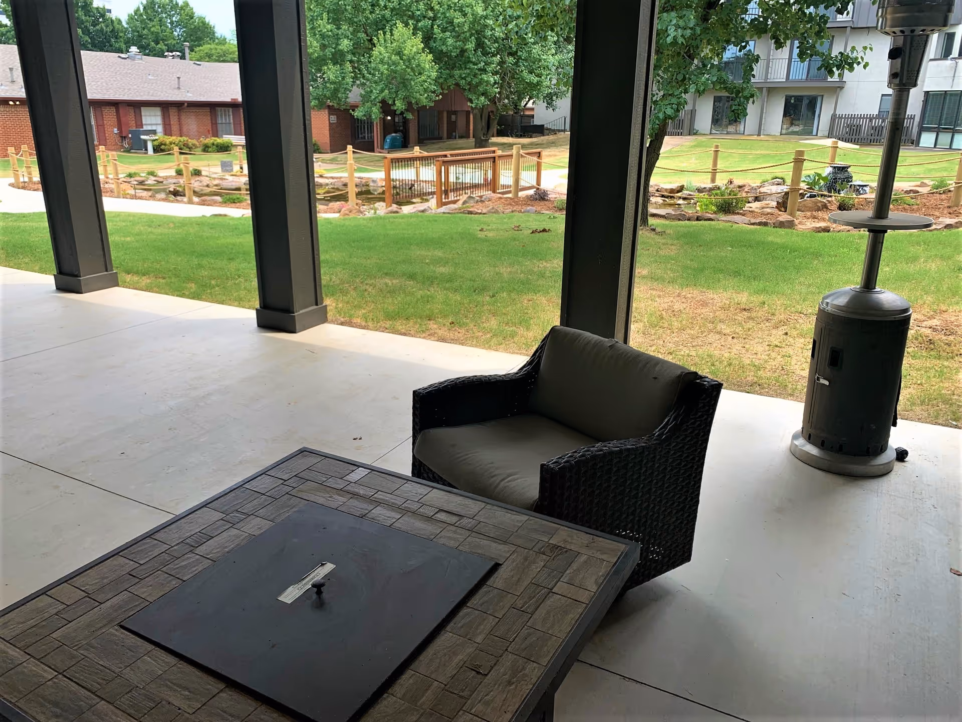 Covered patio with a wicker chair, table and patio heater overlooking a grassy courtyard and neighboring buildings.