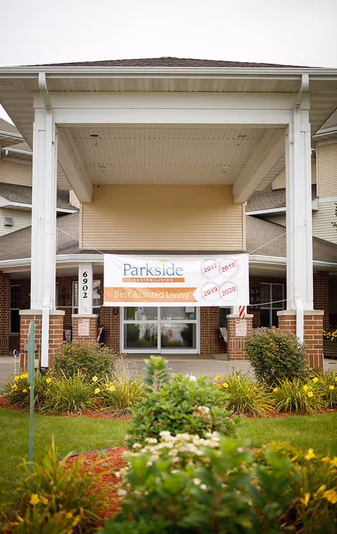 Entrance of Parkside Senior Living facility with a covered porch supported by white pillars. A banner hangs at the entrance stating 'Parkside Senior Living Best Assisted Living' with years 2017, 2018, 2019, and 2020 marked on it. The building has beige siding and brick accents, surrounded by green shrubs and yellow flowers.