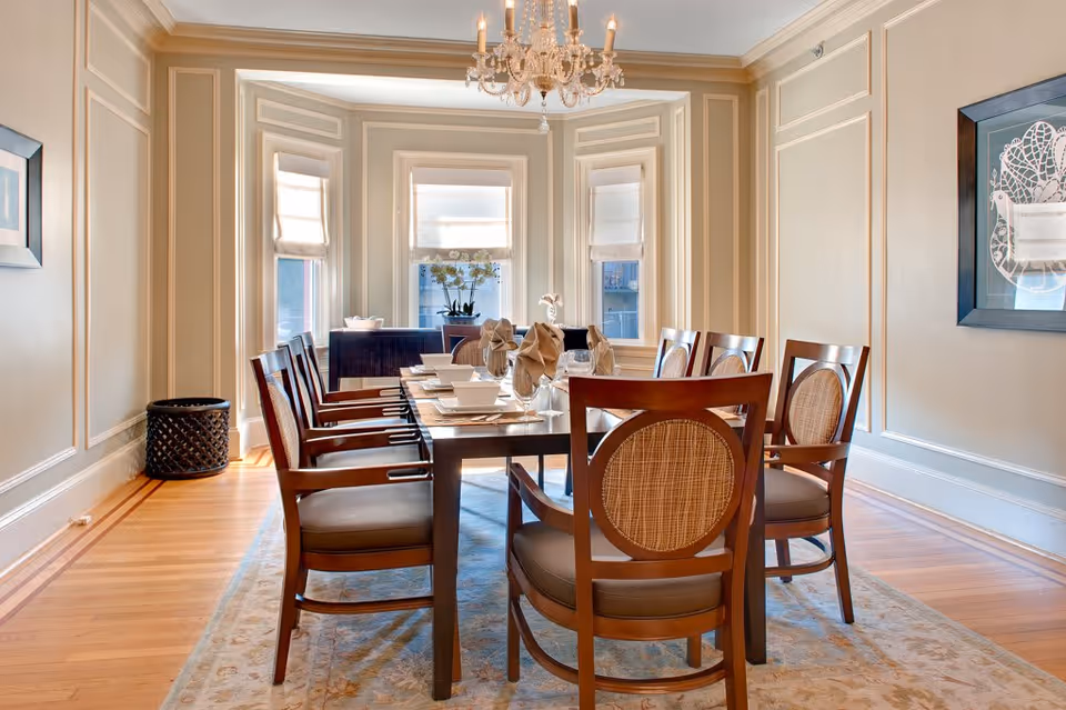 A well-lit dining room with a rectangular wooden dining table set for eight. The chairs have wooden frames with cushioned seats and woven backs. The room features three tall windows with white shades, beige walls with decorative molding, a chandelier hanging from the ceiling, and a light-colored area rug under the table. There is a framed artwork on the right wall and a decorative basket in the corner.