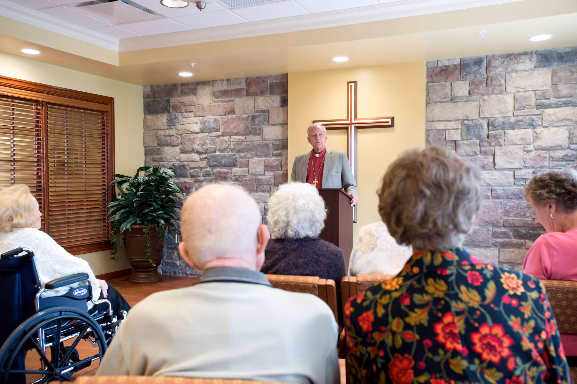 A group of elderly people seated and listening to a man speaking at a podium in a room with stone accent walls and a large wooden cross mounted on the wall behind him. One person is in a wheelchair, and the setting appears to be a chapel or meeting room in a senior living facility.
