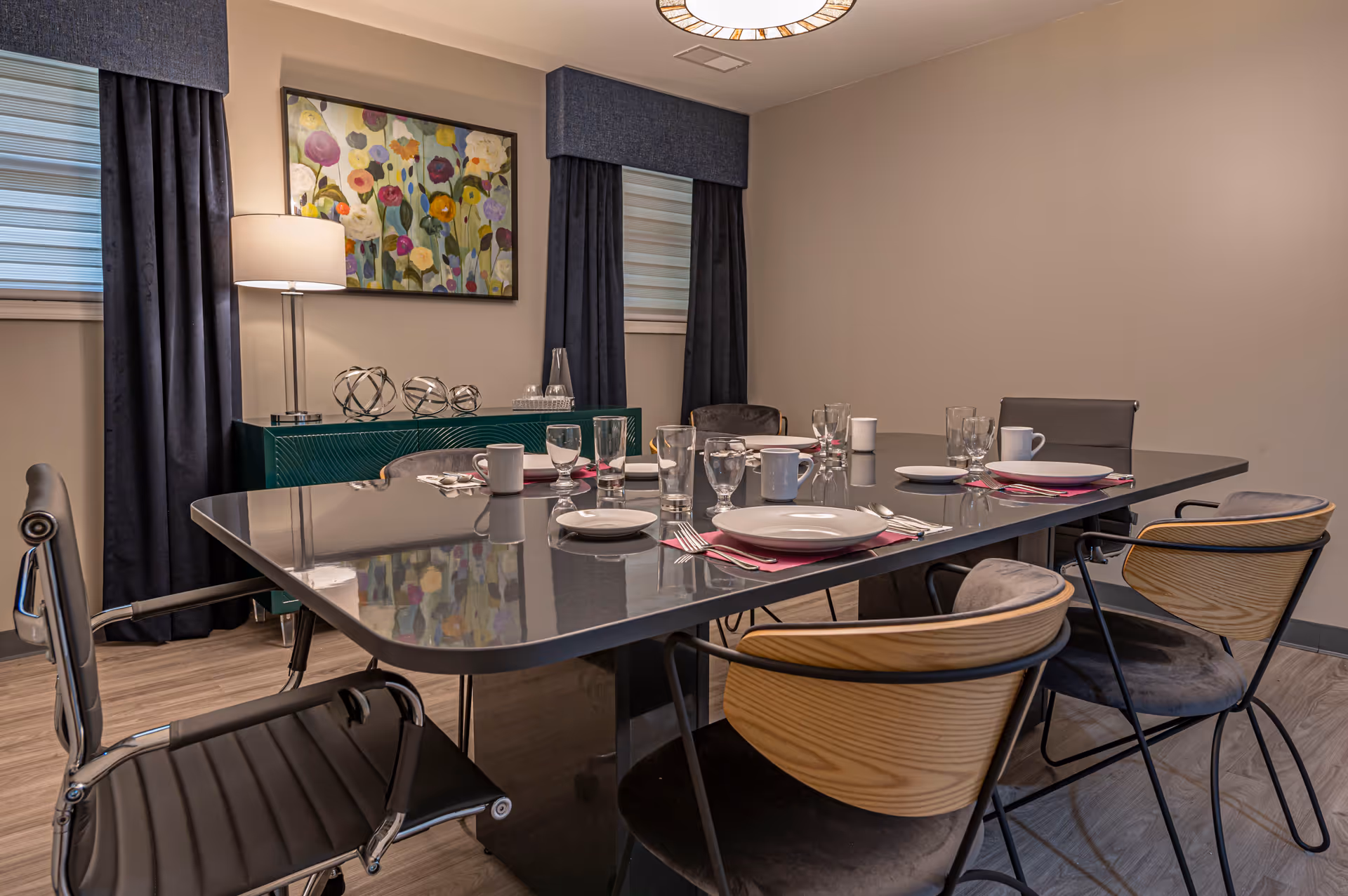 A modern dining room with a rectangular black table set for six people with plates, glasses, cups, and silverware on red napkins. The room has wooden flooring, two windows with dark curtains, a floor lamp, a green sideboard with decorative items, and a colorful floral painting on the wall.