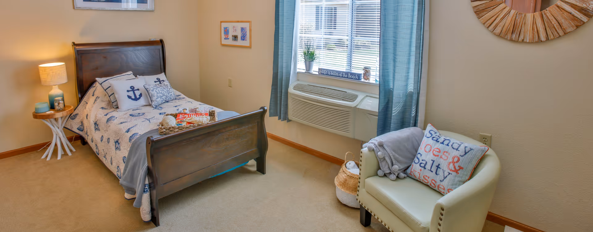 A cozy bedroom with a wooden bed featuring nautical-themed bedding and pillows. Next to the bed is a small round side table with a lamp and decorative items. A window with blue curtains and a window air conditioning unit is on the wall. In front of the window is a light-colored armchair with a pillow that reads 'Sandy toes & salty kisses' and a folded blanket. A woven basket is placed beside the chair, and a decorative round mirror hangs on the wall.