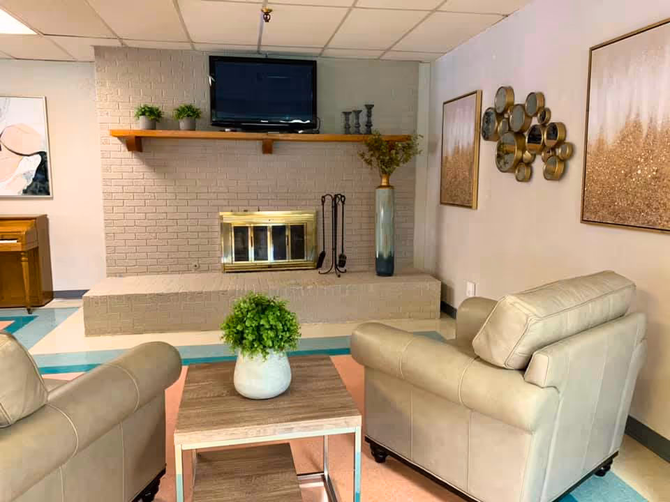 Cozy sitting area with two beige armchairs facing a brick fireplace topped by a wall-mounted TV and a coffee table with a potted plant.
