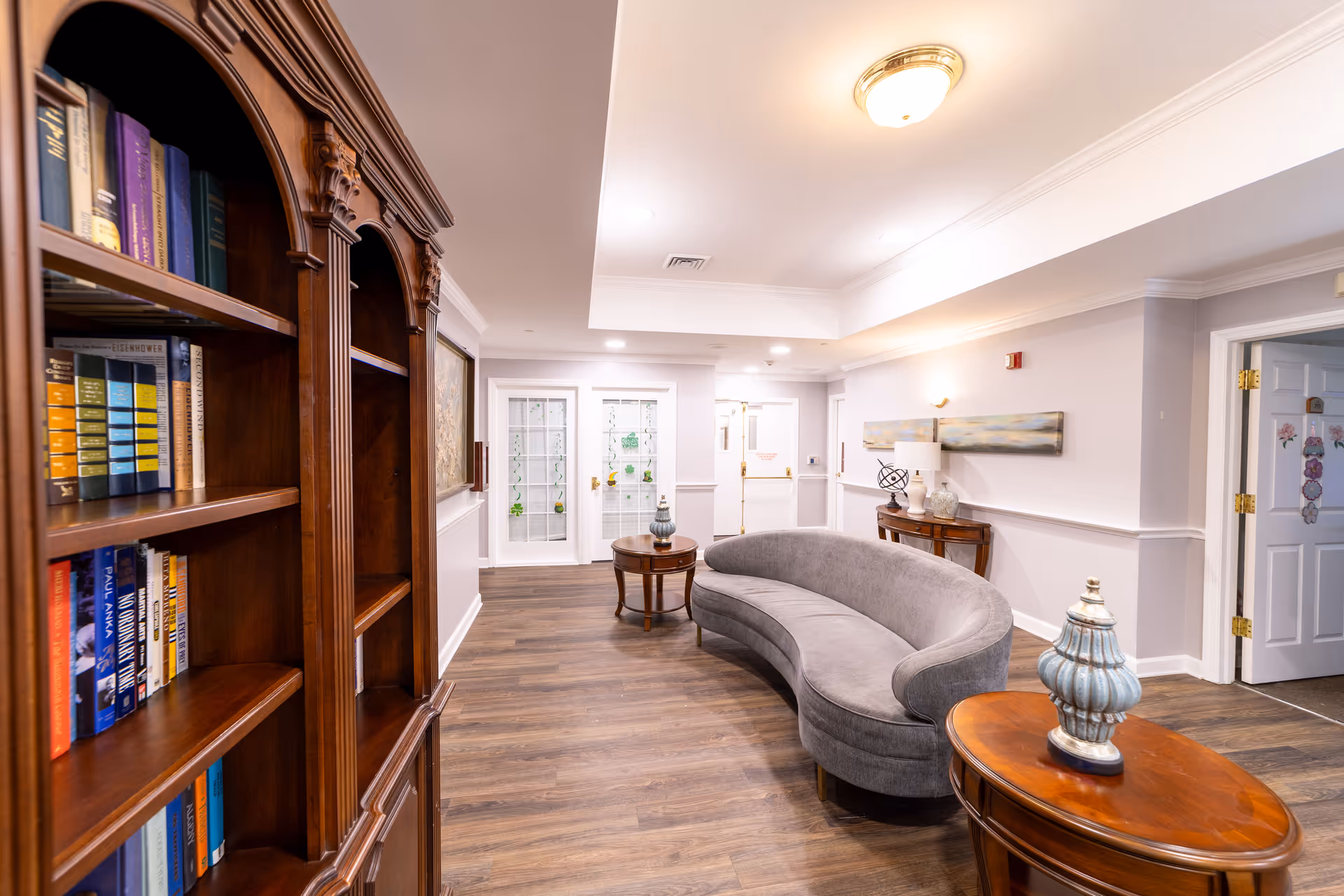 Well-lit lounge in a senior living facility with a curved gray sofa, wooden side tables, and a bookcase along a bright hallway.
