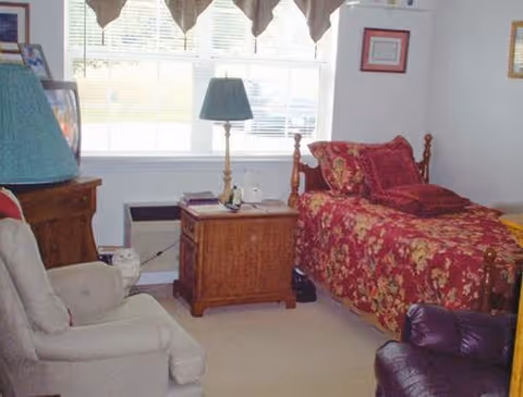 A cozy bedroom with a single wooden bed covered in a red floral bedspread and matching pillows. Next to the bed is a wooden nightstand with a lamp and some items on it. There is a large window with valance curtains letting in natural light. The room also contains a beige armchair and a dark purple recliner chair.