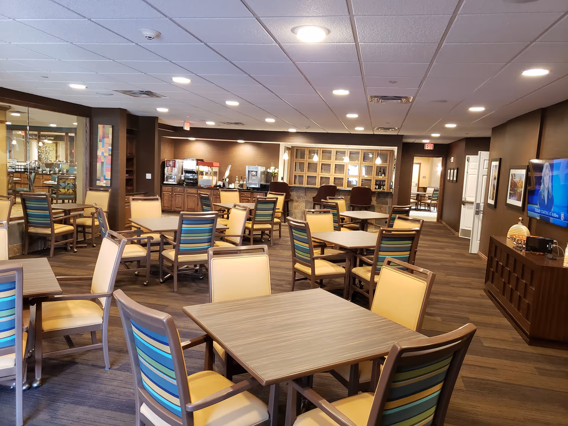 A spacious dining room in a retirement community with multiple tables and chairs arranged neatly. The chairs have striped backs in shades of blue and green with beige seats. The room features a coffee and beverage station along the back wall, warm lighting, and a television mounted on the right wall. The floor is wooden, and the ceiling has recessed lighting.