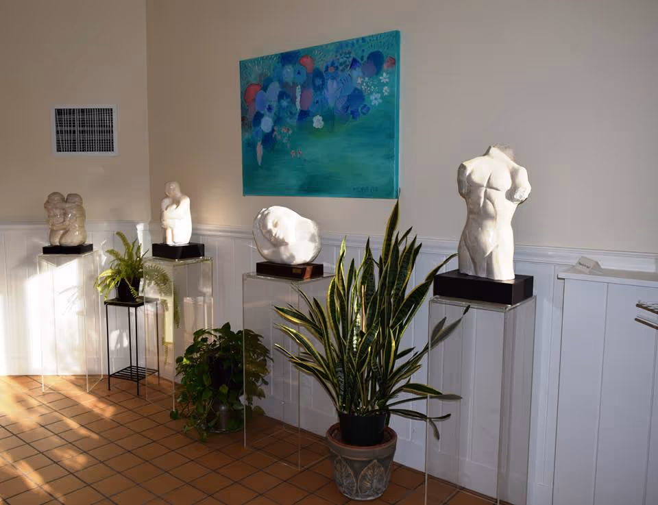 A corner of a room featuring four white sculptures displayed on clear pedestals, with two potted plants placed on the floor and a small black stand. A colorful abstract painting with blue and green tones hangs on the wall above the sculptures. The floor is tiled with a warm brown color, and the walls are painted light beige with white wainscoting.