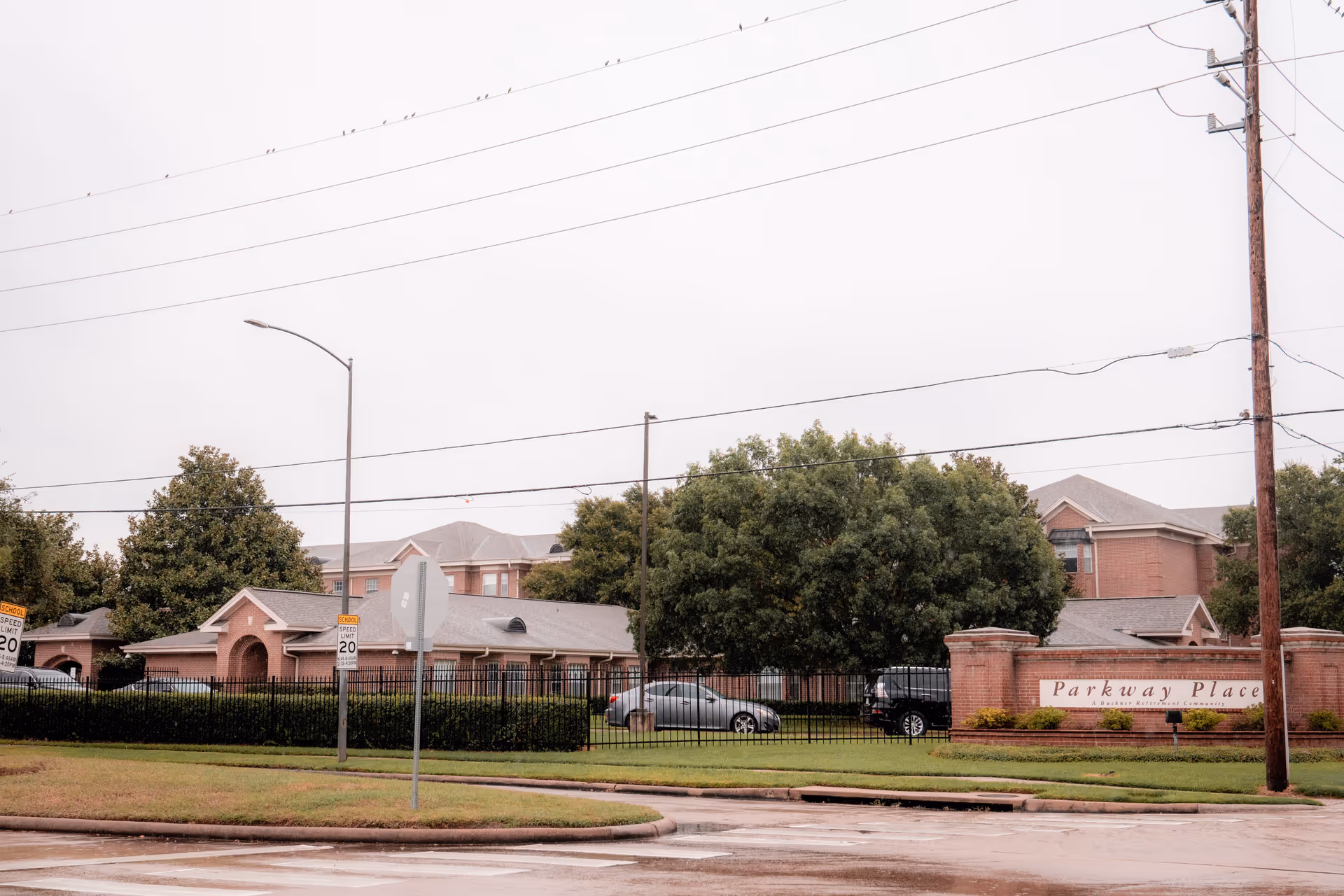 Exterior view of Buckner Parkway Place, a retirement community with brick buildings, green trees, a black metal fence, and a sign reading 'Parkway Place A Buckner Retirement Community' near a street corner with traffic signs and power lines overhead.