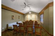 Interior view of a small chapel or meditation room with rows of wooden chairs facing a cross mounted on the far wall. The room has a high ceiling with ceiling fans, beige walls, a carpeted floor, and a window on the right side letting in natural light.