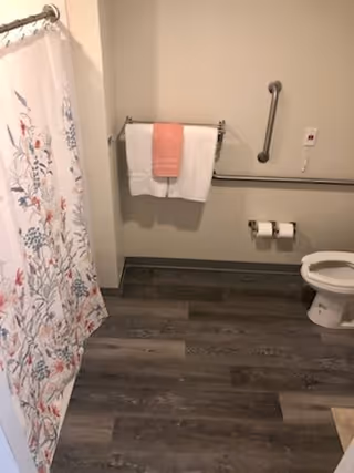 A bathroom with a floral-patterned shower curtain on the left, a towel rack with a white towel and a pink towel hanging on the wall, a grab bar, two toilet paper rolls on a holder, and a white toilet on the right. The floor has dark wood-like flooring.