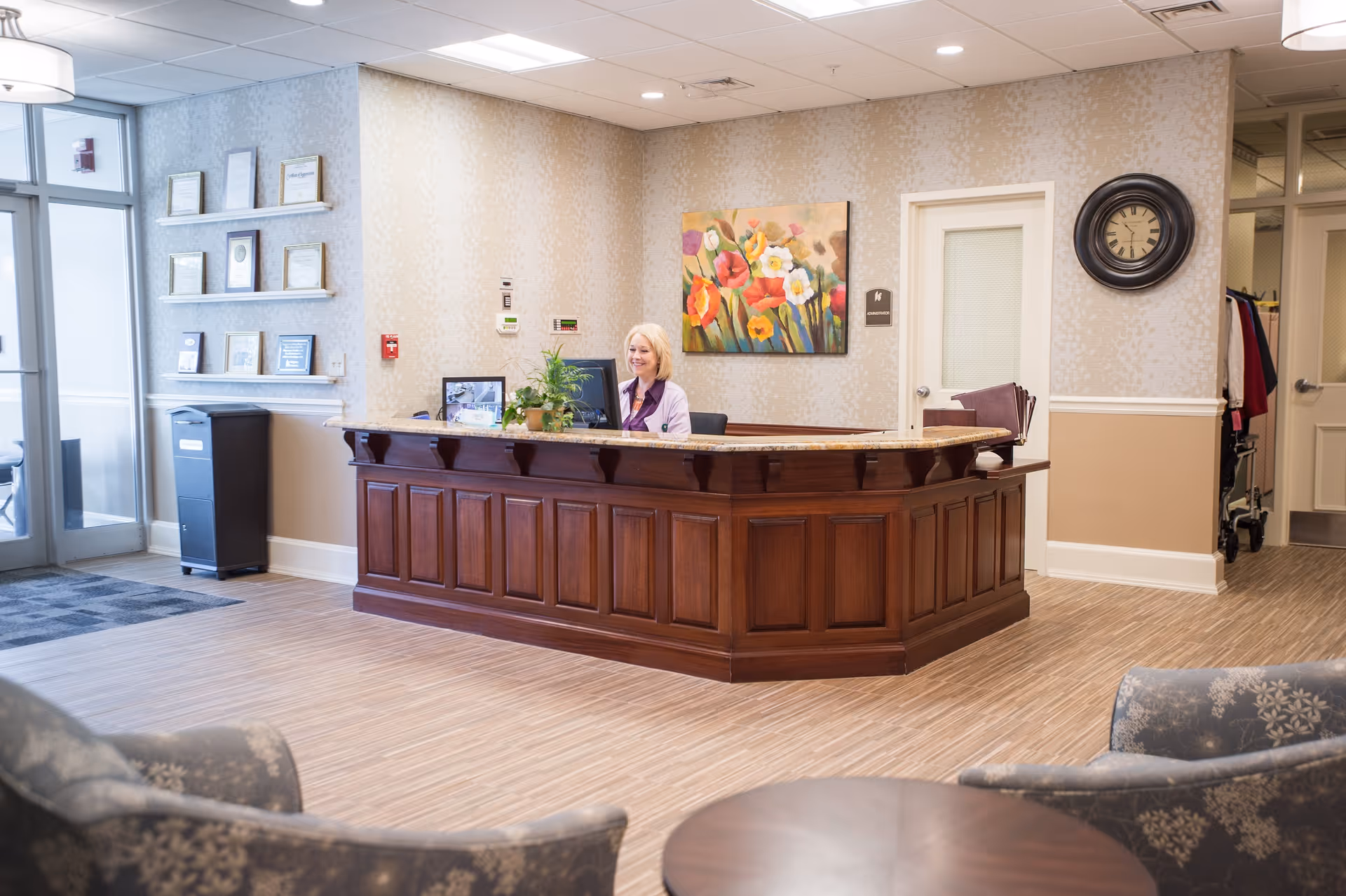 Reception area of Kingsway Arms Nursing Center with a wooden front desk, a receptionist sitting behind the desk, a colorful floral painting on the wall, a round wall clock, and comfortable seating in the foreground.