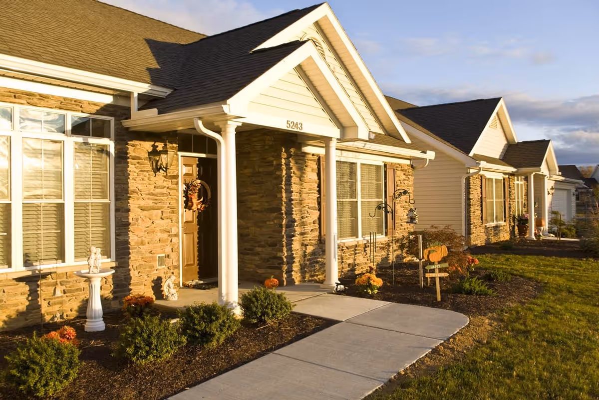 Exterior view of a residential building with stone and siding facade, a brown front door decorated with a wreath, white columns supporting a small porch roof, and a concrete walkway leading to the entrance. The landscaping includes bushes, flowers, and small garden decorations, with a lawn and partly cloudy sky in the background.