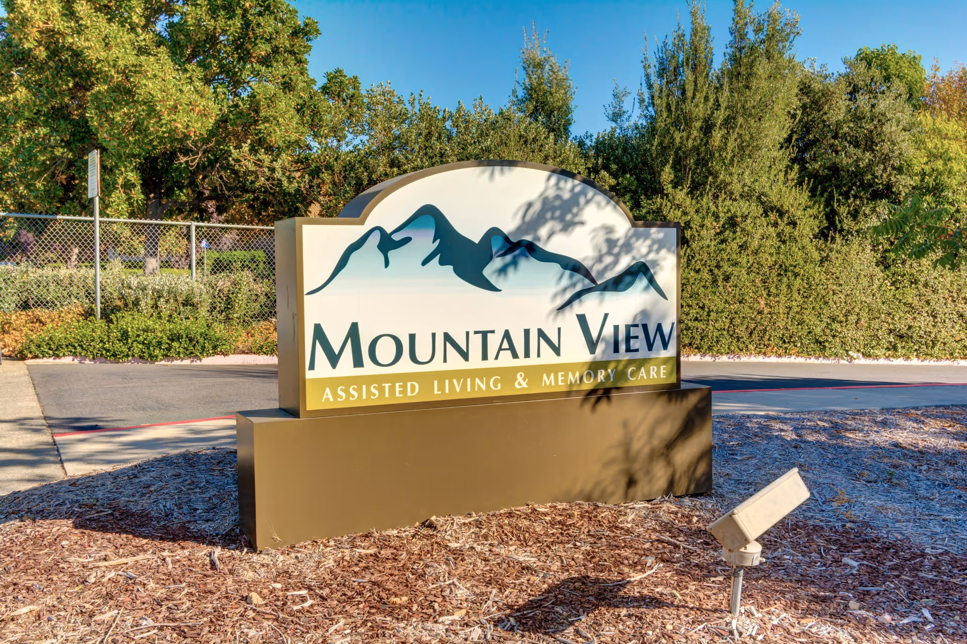 Outdoor sign for Mountain View Assisted Living & Memory Care facility, surrounded by mulch and greenery with trees and a chain-link fence in the background under a clear blue sky.