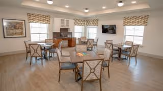 A bright and spacious dining area with several wooden tables and chairs arranged neatly. The room has large windows with patterned valances allowing natural light to fill the space. There is a wall-mounted television and cabinetry with a countertop along one wall.