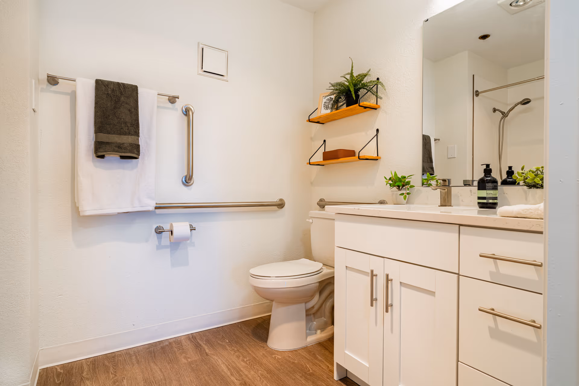 A clean and modern bathroom with a white toilet, white vanity with a sink, and a large mirror above the sink. There are two wooden shelves on the wall holding a small plant, a framed picture, and a basket. A towel rack with a white and a dark gray towel is mounted on the wall next to the toilet, along with two stainless steel grab bars. The floor has a wood-like finish.