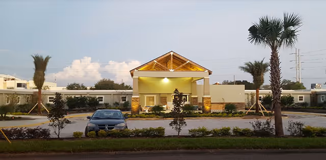 Front exterior of a single-story assisted living building with a covered entrance, palm trees, landscaping, and a parked car in front.