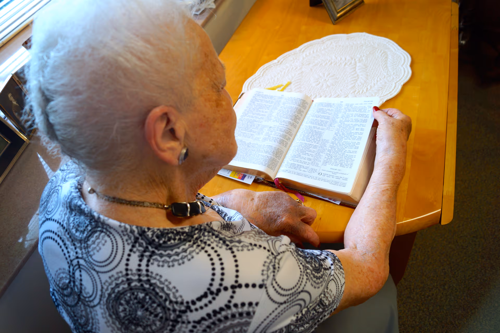 An elderly woman with white hair is sitting at a wooden table reading an open book. She is wearing a patterned shirt and a necklace. The table has a white lace doily and some other small items on it. The setting appears to be indoors near a window with natural light coming in.