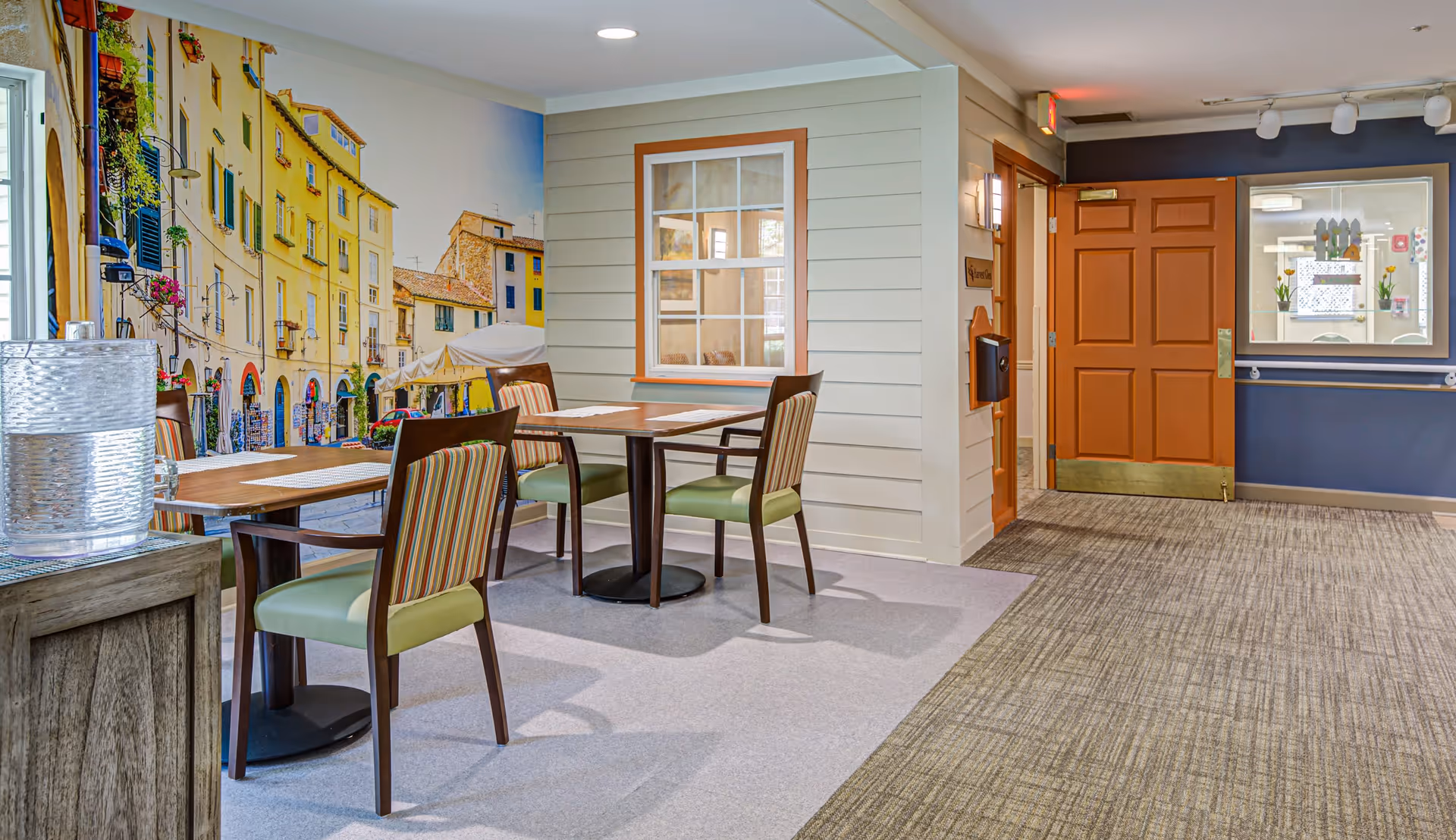 Interior view of a dining area in a senior living facility with two wooden tables and four chairs with green cushions and striped backs. A large wall mural depicts a colorful street scene with yellow buildings and flowers. There is a wooden side table with a textured glass water dispenser on the left. The floor is carpeted with two different patterns, and a wooden door with a brass kick plate is visible in the background along with a window displaying decorations.