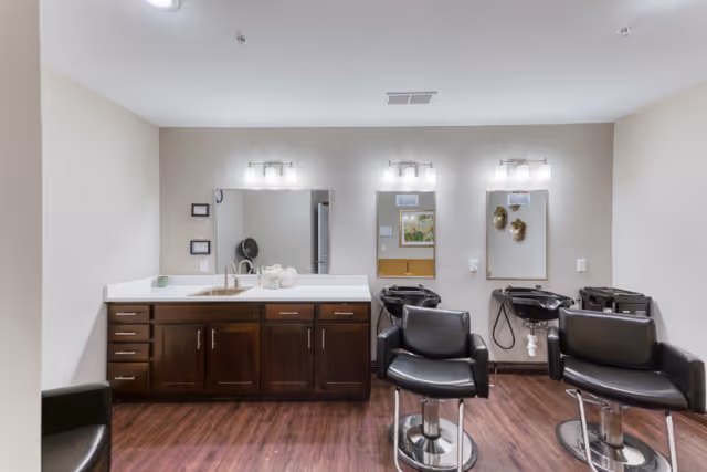Interior view of a salon area in a senior living facility with two black salon chairs in front of two hair washing stations with sinks and mirrors. To the left, there is a countertop with a sink, cabinets, and three mirrors above. The floor is wooden, and the walls are painted light beige.