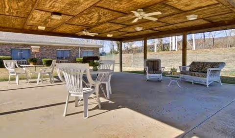 Covered outdoor patio area with white plastic chairs and tables, wicker armchairs, and a wicker sofa with floral cushions. Ceiling fans are mounted on the wooden ceiling. In the background, there is a brick building and a grassy area with trees.