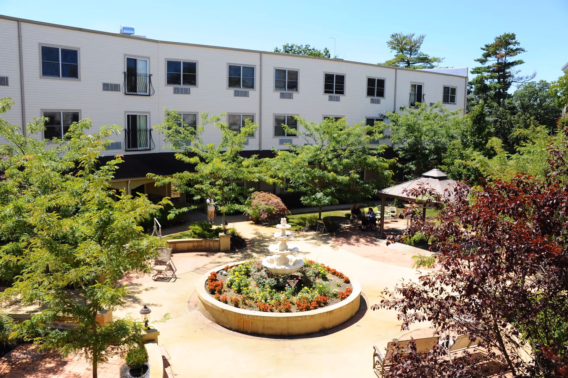 Outdoor courtyard area at Lakewood Courtyard featuring a circular flower bed with a tiered fountain in the center, surrounded by trees, benches, and a gazebo with seating. The background shows a multi-story building with windows and balconies under a clear blue sky.