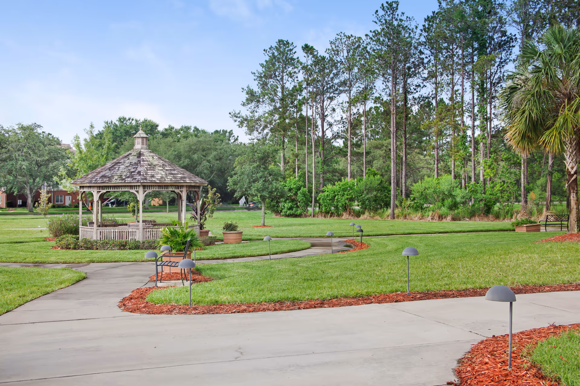 A well-maintained outdoor garden area featuring a wooden gazebo surrounded by green grass, trees, and paved walkways with small pathway lights. There are benches and potted plants along the paths under a clear blue sky.