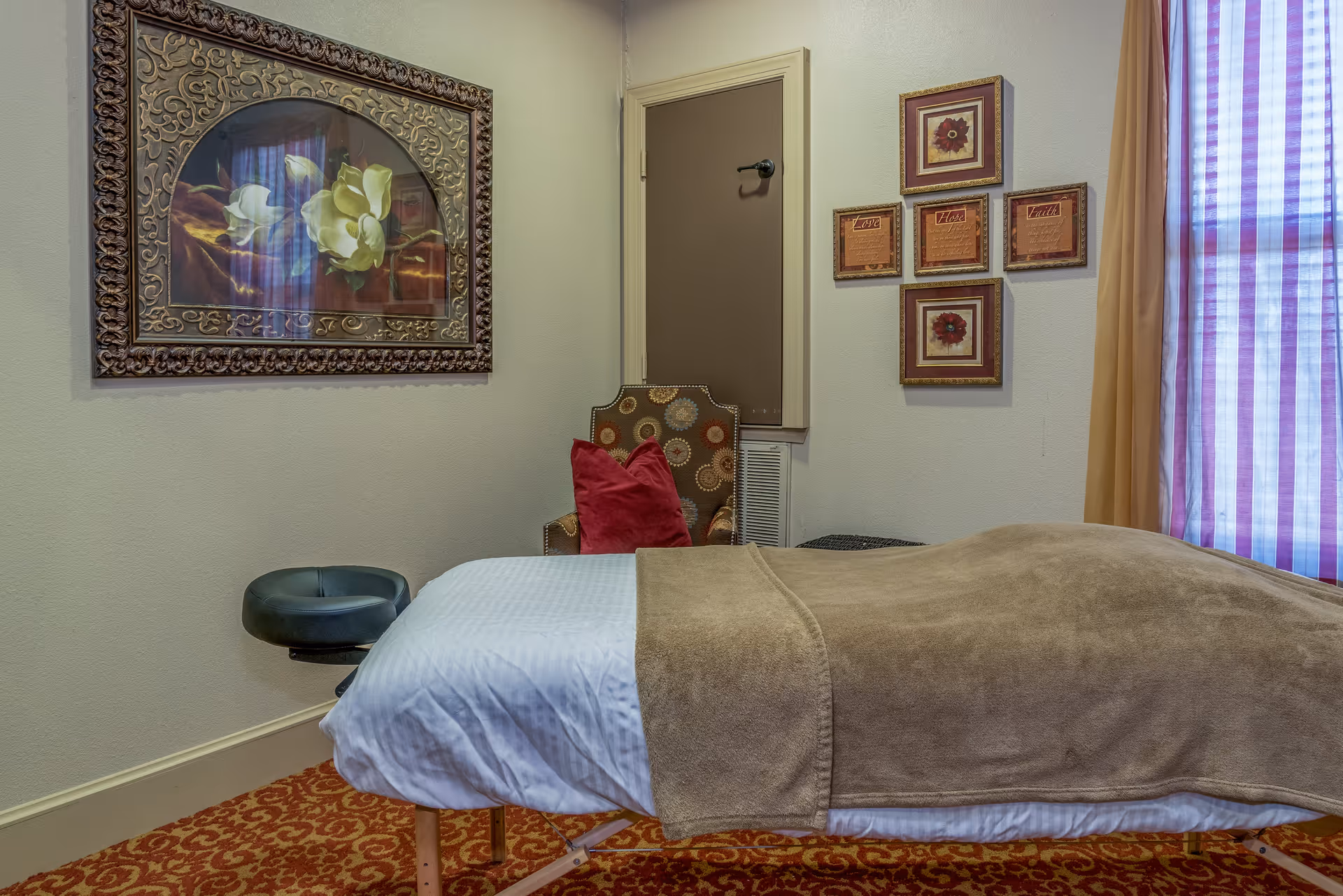 A cozy room with a massage table covered with a white sheet and a brown blanket. There is a patterned armchair with a red pillow in the corner, a framed floral painting on the wall, and a window with sheer curtains letting in light. The room has beige walls and a red patterned carpet.
