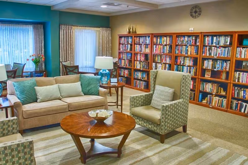A cozy living room area in a senior living facility with a beige sofa adorned with green and beige cushions, a patterned armchair with a matching cushion, a round wooden coffee table with a decorative bowl, and a side table with a blue lamp. Behind the seating area is a large wooden bookshelf filled with books. The room has large windows with sheer curtains and a green accent wall.