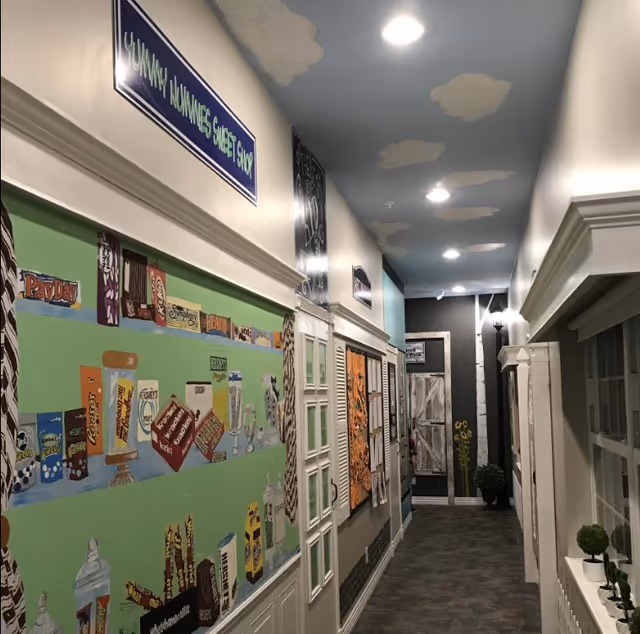 Interior hallway decorated with colorful murals, faux storefront doors and a cloud-painted ceiling.