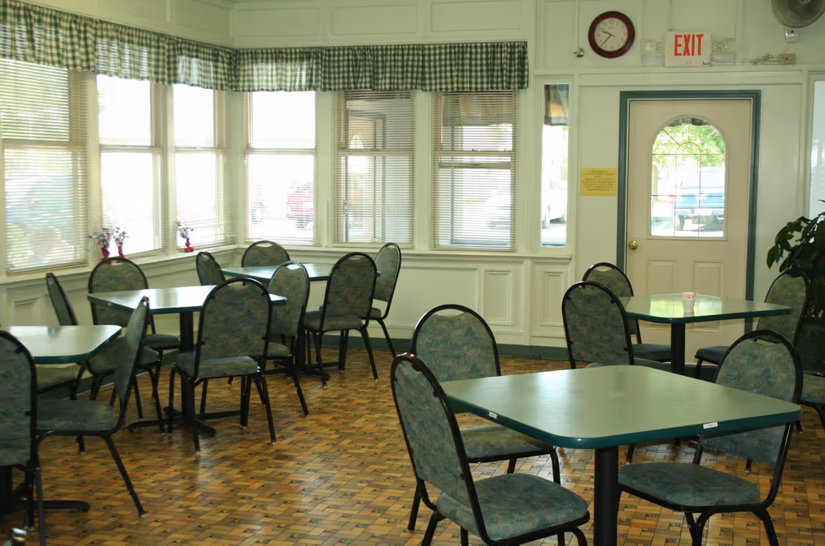 Small dining room with green tables and padded chairs, large windows and an exit door.