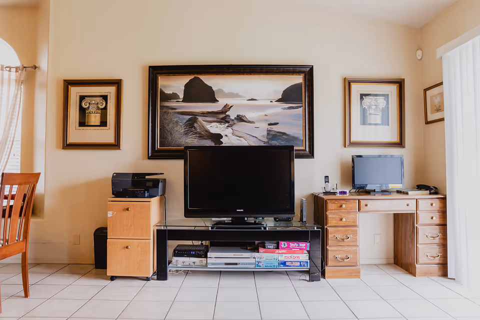 A living room area featuring a flat-screen TV on a glass and metal stand with board games and electronic devices underneath. To the left is a wooden cabinet with a printer on top, and to the right is a wooden desk with a computer monitor, phone, and office supplies. The walls are decorated with framed artwork, including a large landscape painting above the TV. The floor is tiled, and there is a wooden chair partially visible on the left side.