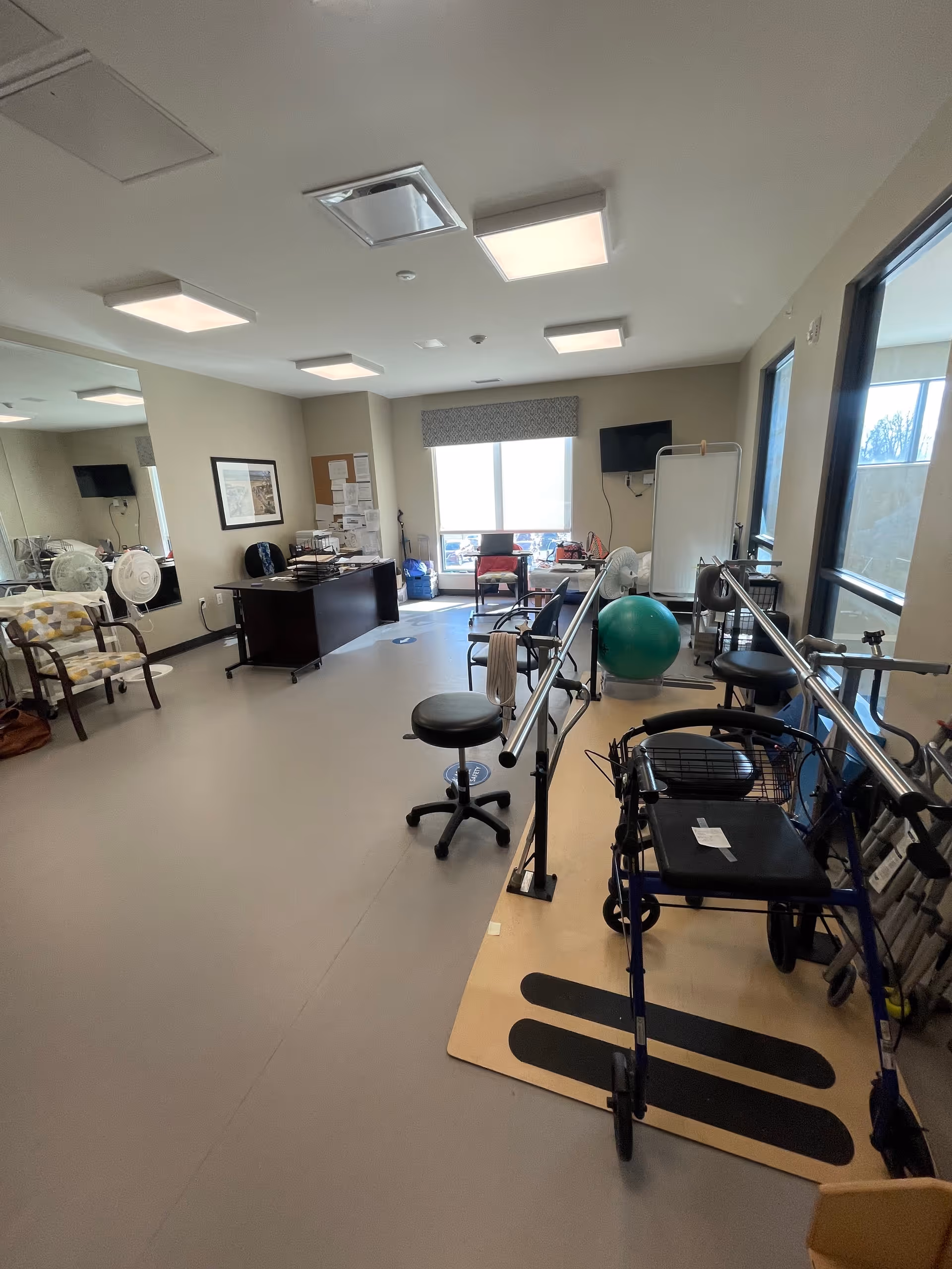 A rehabilitation or physical therapy room with parallel bars for walking exercises, a walker, a green exercise ball, stools, chairs, a desk with office supplies, a whiteboard, and a wall-mounted TV. Large windows provide natural light.