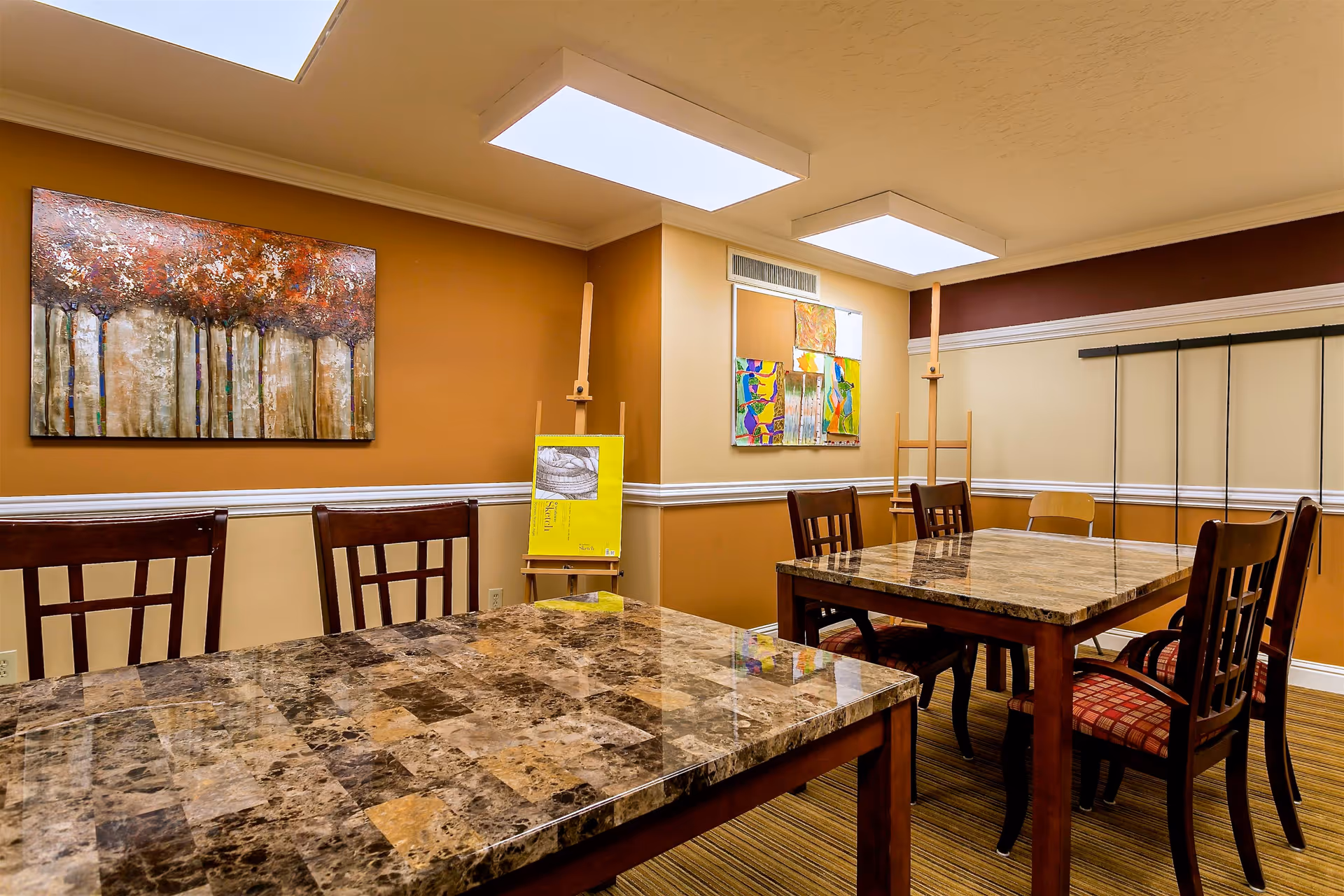 Interior room with two marble-patterned tables surrounded by wooden chairs with red cushioned seats. The walls are painted in warm tones of brown and beige with white trim. The room is decorated with colorful paintings and two easels, and has two rectangular ceiling lights.