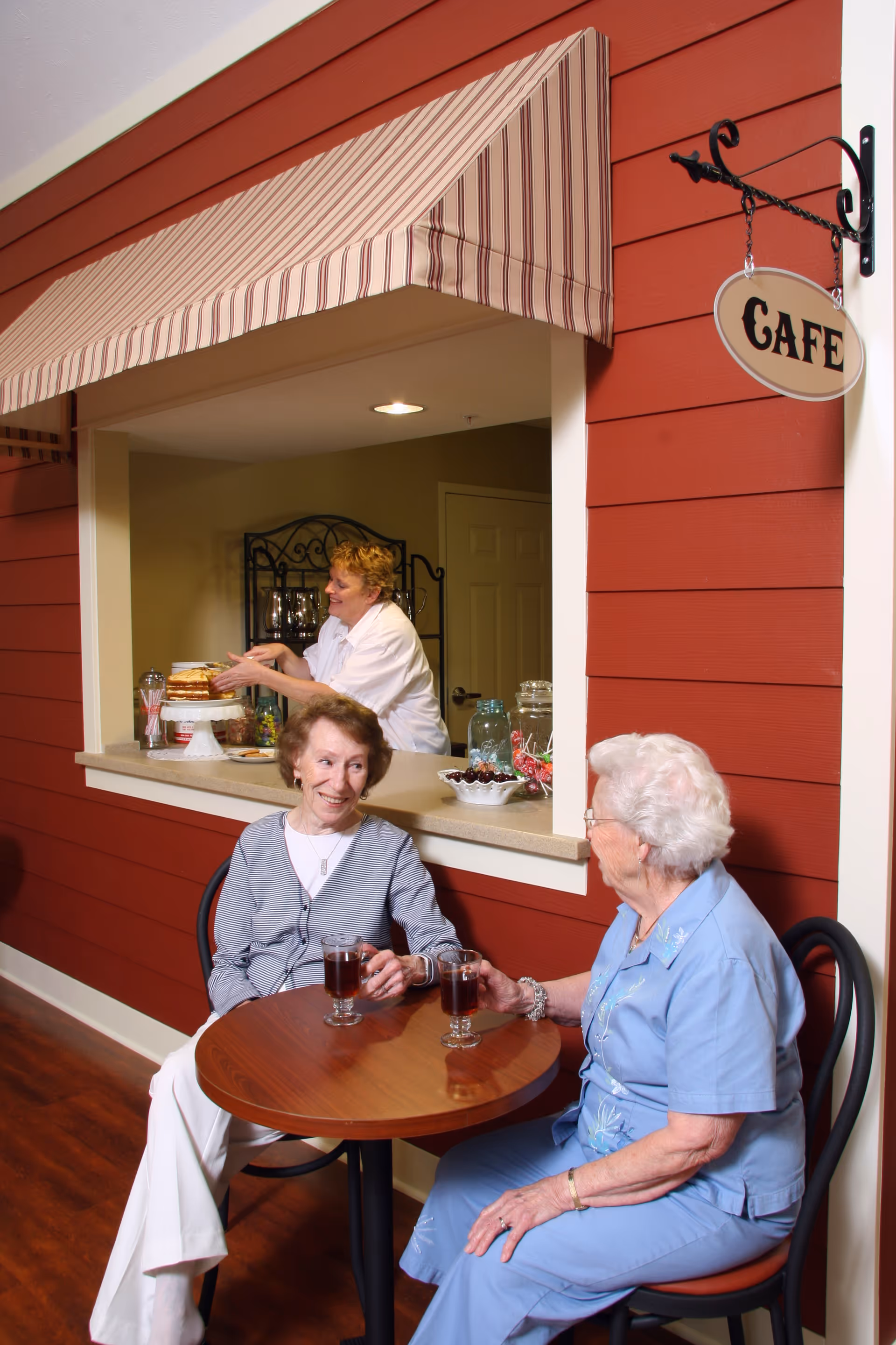 Two elderly women sitting at a small round wooden table with drinks, chatting and smiling in front of a cafe window where a woman is serving food. The cafe window has a striped awning and a hanging sign that says 'CAFE'. The setting has red paneled walls and wooden flooring.