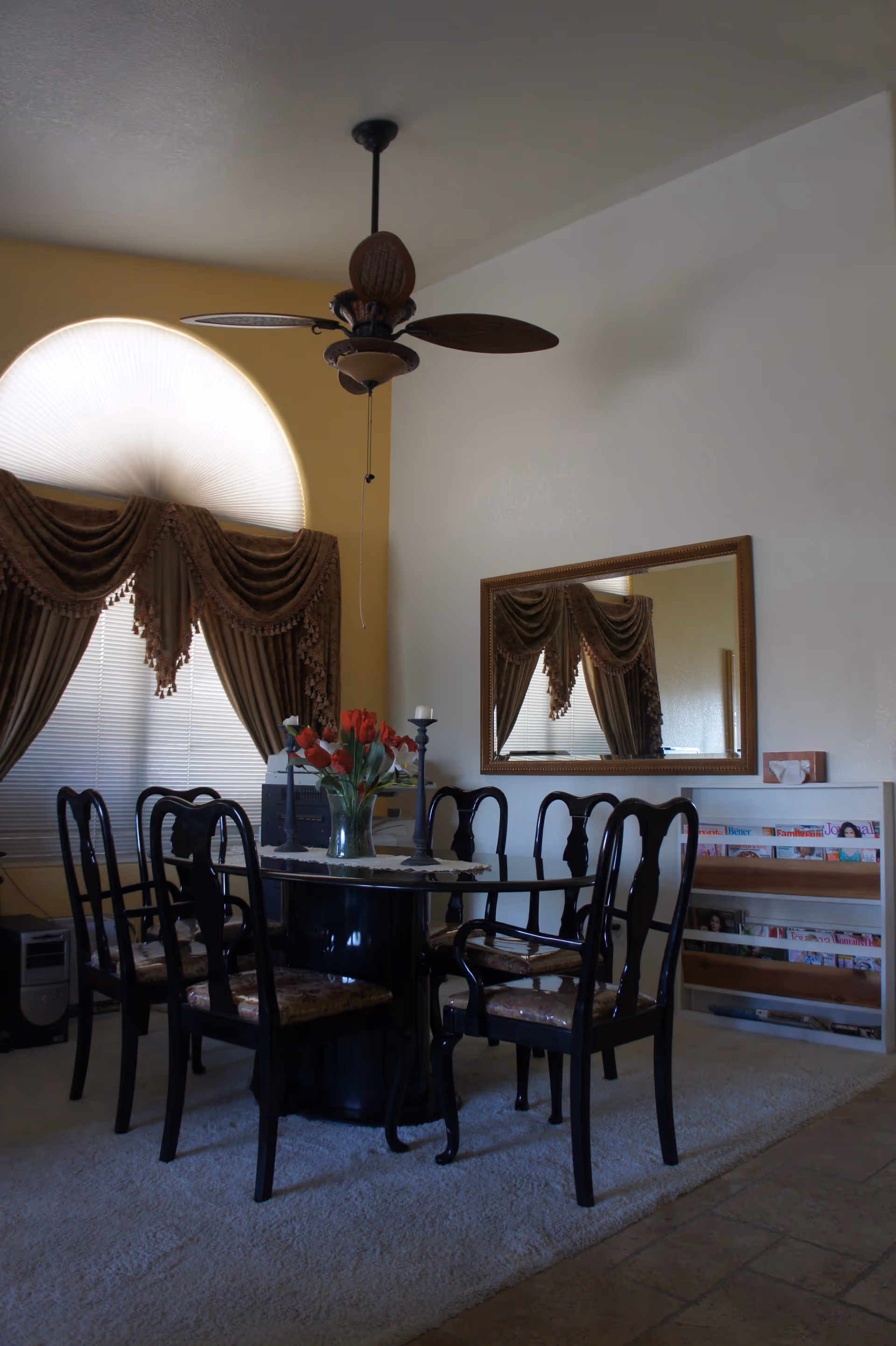 A dining room with a round black table surrounded by six black chairs with patterned cushions. A vase with red flowers and two candle holders are on the table. The room has a large arched window with brown drapes and blinds. A ceiling fan with wooden blades hangs above the table. A large rectangular mirror is mounted on the wall, reflecting the window and drapes. A white shelf with magazines is against the wall.
