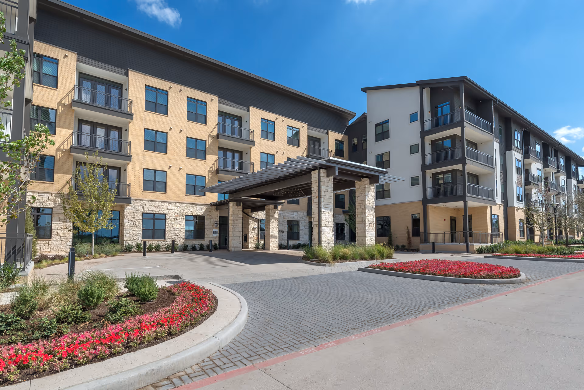 Exterior view of a modern multi-story senior living facility with balconies, a covered entrance, landscaped flower beds, and a clear blue sky.