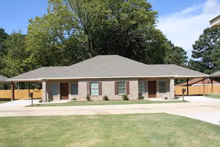 Single-story brick building with a gray shingled roof, multiple doors and windows, surrounded by a concrete driveway and green lawn, with trees in the background under a blue sky.