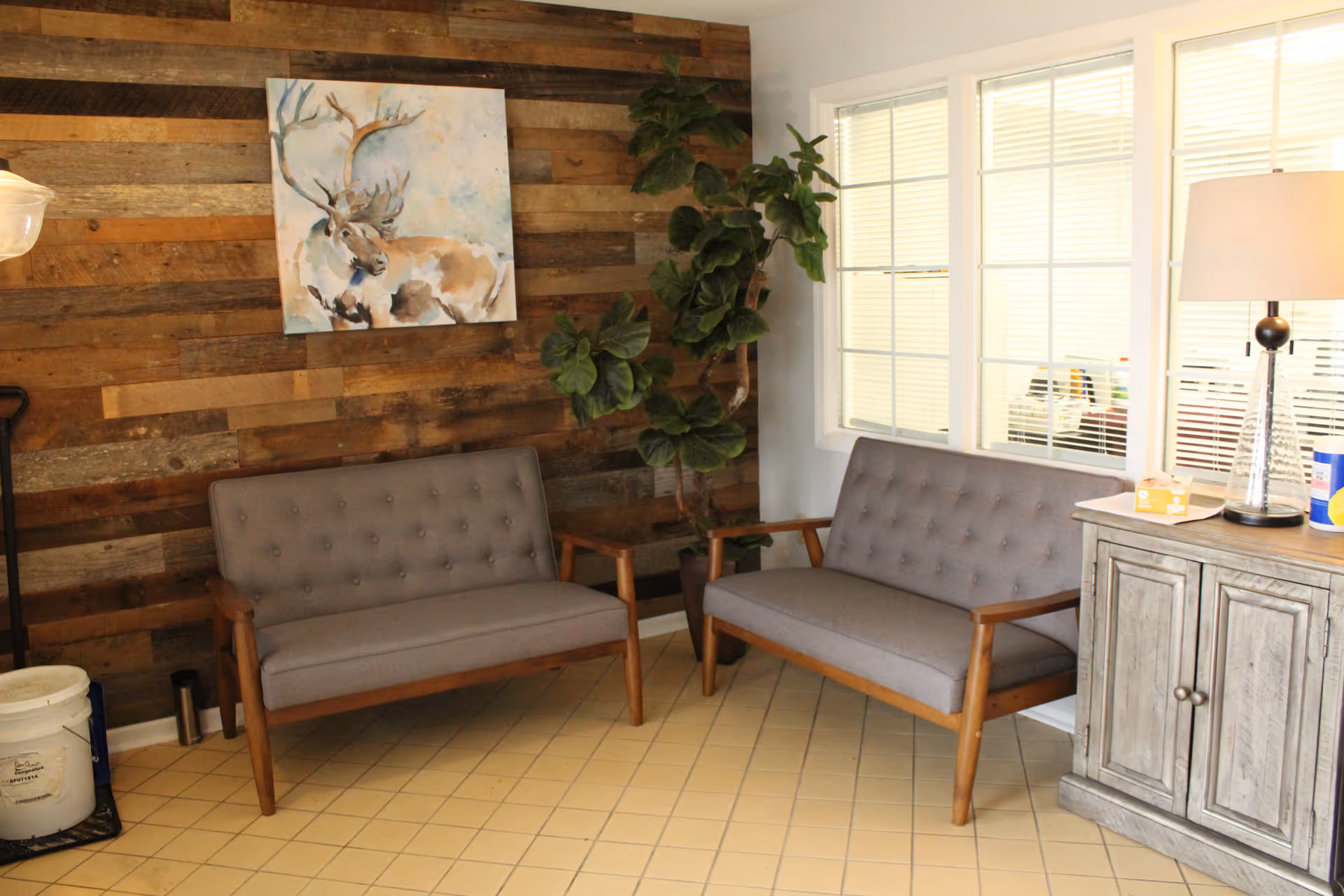 A cozy seating area with two gray cushioned wooden armchairs placed against a rustic wooden plank wall. A painting of a moose hangs on the wall above one chair. To the right, there is a window with white blinds and a wooden cabinet topped with a clear glass lamp and a box of tissues. A tall green potted plant stands between the chairs and the window.