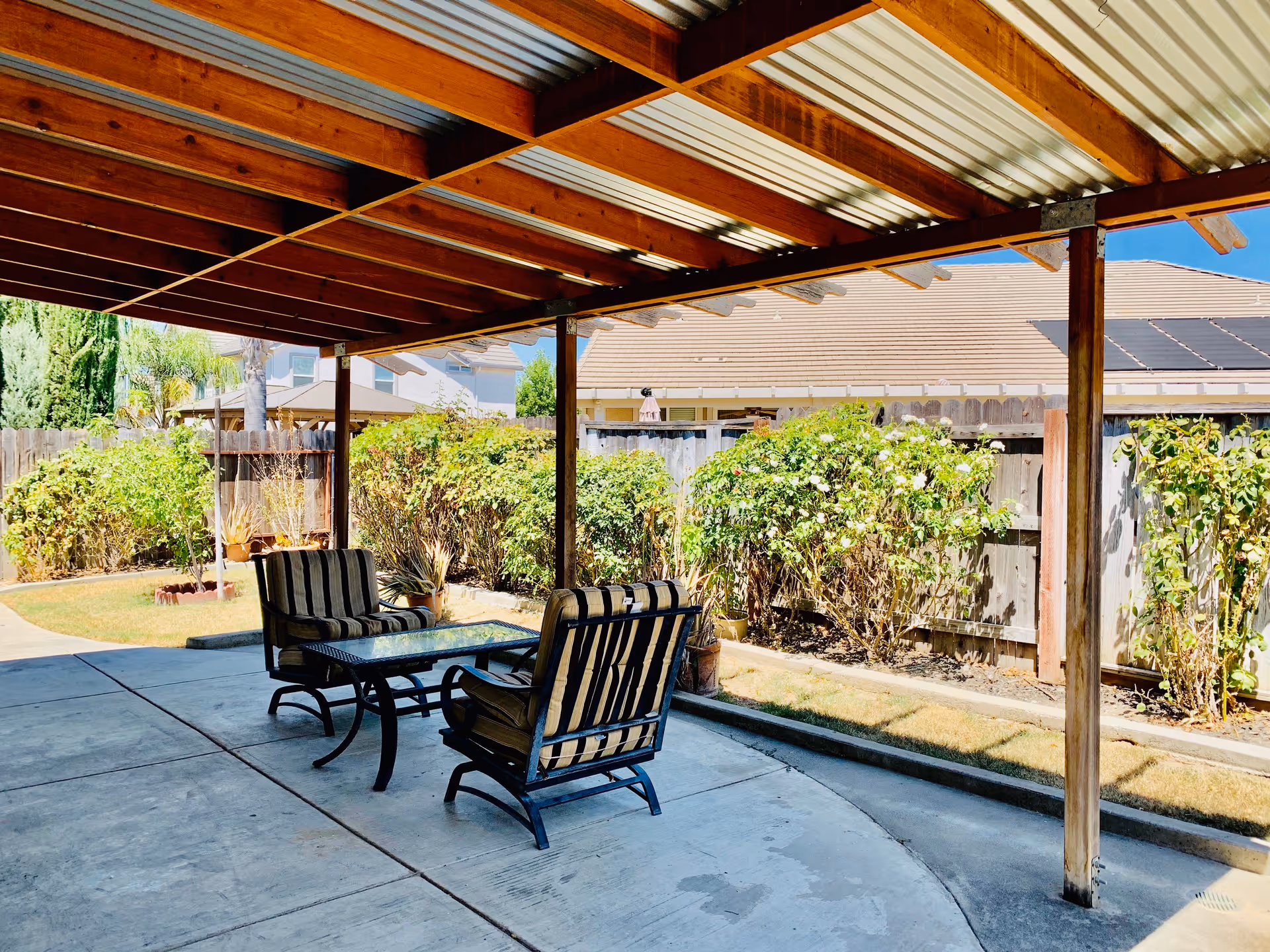 Outdoor patio area with a wooden pergola roof, two cushioned chairs, and a glass-top table on a concrete floor. The patio overlooks a garden with bushes and a wooden fence, with neighboring houses visible in the background under a clear blue sky.