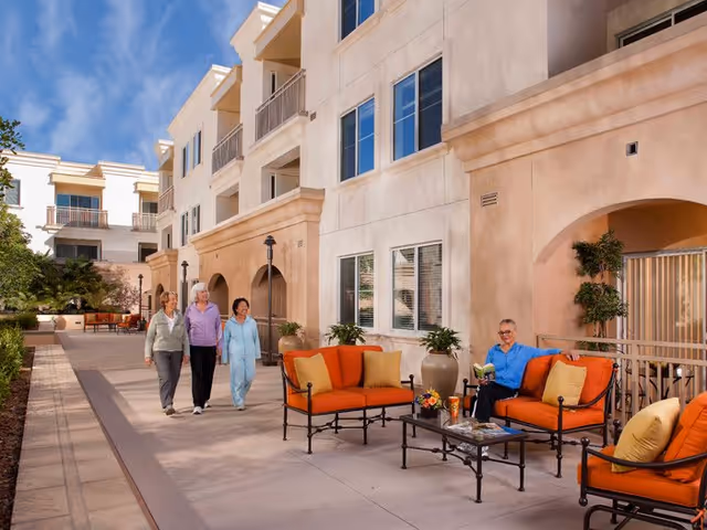 Outdoor patio area at a senior living facility with beige building walls, orange cushioned seating, potted plants, and four elderly women, three walking together and one sitting and reading a book.