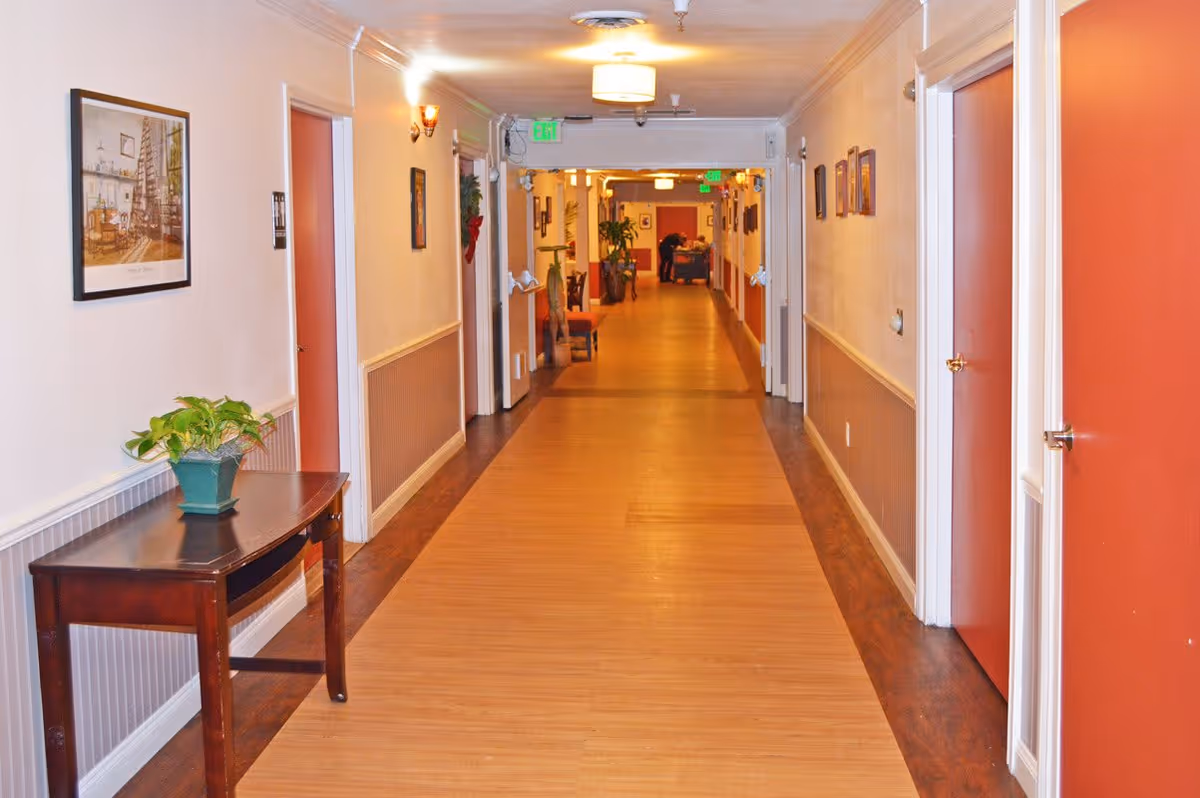 Long, well-lit interior hallway with wood floors, doors on both sides, a small table with a potted plant, and seating visible at the far end.