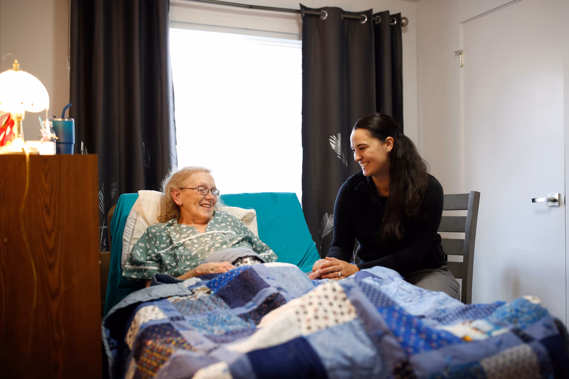 An elderly woman lies in a bed covered by a blue patchwork quilt, smiling as a caregiver sits beside her.
