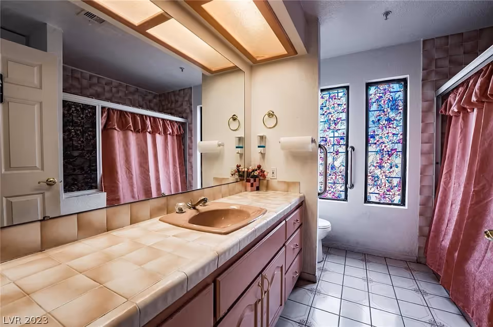 Bathroom with a long tiled countertop and a pink sink, large mirror above the sink, pink curtains on the shower, and two tall windows with colorful stained glass. The floor is tiled with a white and gray pattern.