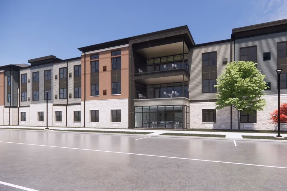 Exterior view of a modern three-story senior living facility building with large windows, balconies, and a parking area in front. There are trees and street lamps along the sidewalk.